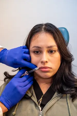 A woman wearing a nose ring and a zipper jacket has her nose pierced by a doctor wearing gloves and holding a syringe.