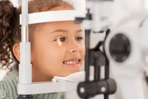 A young girl with curly hair is having her eyes examined with a slit lamp in a medical setting.
