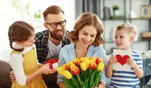 A family, including a woman, a man, and two children, is sitting on a couch, smiling and holding a bouquet of tulips and a gift
