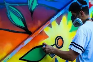 A man wearing a mask and a white shirt spray-painting a colorful mural on a wall