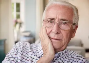 An elderly man wearing glasses and a plaid shirt is looking down with a hand on his chin.