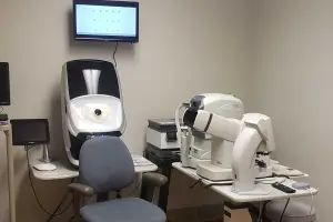 A medical examination room with various eye care equipment on white desks and a mounted monitor on the wall.