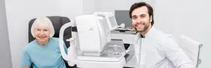 A woman is sitting in front of an eye exam machine with a smiling man standing behind her in a white coat.