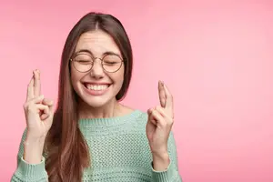 A woman with glasses, smiling and making a peace sign with her fingers in front of a pink background