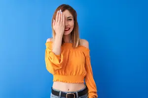 A woman covering her face with her hand while standing in front of a blue background.