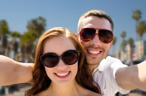 A man and woman smiling and posing for a selfie with sunglasses on a beach with palm trees in the background