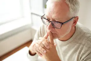 An elderly man with gray hair and glasses wearing a white shirt sitting and looking down with his hand on his chin