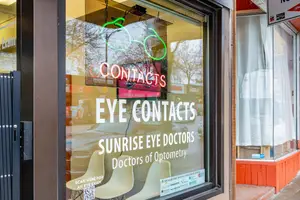 A storefront window displays a neon sign that reads "Contacts Eye Contacts Sunrise Eye Doctors Doctors of Optometry" with a reflection of a car and chairs inside.