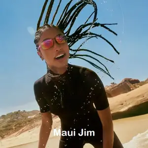 A woman wearing a black wetsuit and sunglasses, smiling with water splashing around her on a beach.