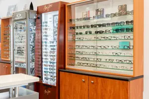 A display of sunglasses in a store with wooden cabinets and glass shelves, featuring various brands and styles of sunglasses, including Oakley, Maui Jim, and others, along with a white table in the foreground.