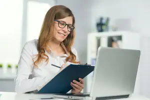 A woman wearing glasses and a white shirt is sitting at a desk, smiling and holding a blue folder with a pen, while looking at a laptop.
