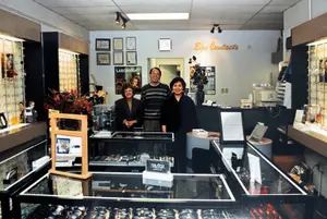 Three people are standing in an office room with a glass counter and display case, smiling and posing for a photo