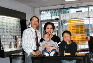 A family of four is smiling for a picture in front of a store with glass doors and windows. The father is wearing glasses and a tie, the mother is wearing glasses and a black top, the baby is in the mother's arms, and the boy is sitting on a counter with his hand on his chin.