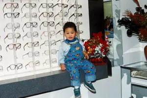 A young boy wearing overalls is sitting on a bench in an optometrist's office with a display of eyeglasses behind him and a floral arrangement in front.