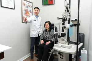 A smiling doctor is standing next to an older woman who is sitting in a chair next to an eye exam machine in an exam room.