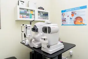 Medical equipment and supplies are displayed in a well-organized room. On the left side, there is a cabinet with various items, including bottles and boxes, neatly arranged. On the right side, a blue poster is attached to the wall, featuring a detailed diagram of the eye's anatomy. In the center, a table holds three eye examination machines, one of which is actively being used, while the other two are positioned in front of it. The room is well-lit, and the overall atmosphere is clean and professional.