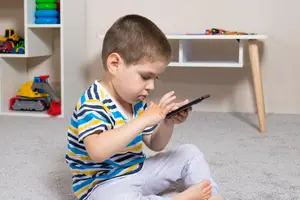 A young boy is sitting on the floor, using a smartphone while surrounded by toys and a white shelf.