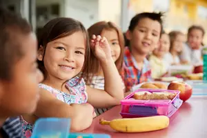Children are sitting at a table with their lunch and are smiling and chatting with each other.