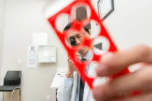 A man in a white lab coat and tie is holding a red ophthalmoscope with multiple circular lenses while looking into a mirror in a medical office.