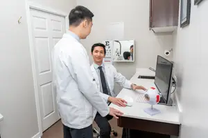 A doctor examines a patient in an eye examination room, with a computer and eye model on the desk, while another doctor looks on.