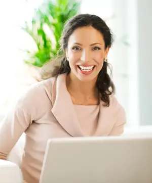 A smiling woman sitting in front of a laptop, wearing a pink sweater and earrings.