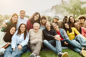 A group of people, including a woman in glasses, a man with a hat, and a young man with a black shirt, are smiling and posing for a photo in a park.