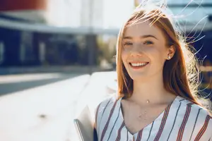 A young woman with red hair wearing a white and red striped blouse and a necklace stands on a sidewalk, smiling and posing for a photo with a book in her hand. Behind her, a blurry background shows a building with glass windows and a street light, suggesting a sunny day with the woman's hair gently blowing in the breeze.