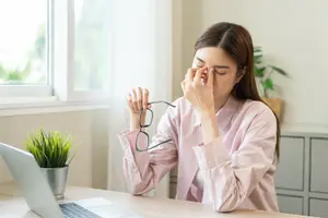A woman sitting at a desk with her eyes closed, holding a pair of glasses and covering her face with her hands.