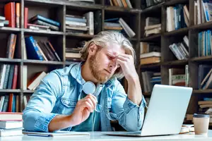Man sitting in front of a desk with a laptop and headphones looking frustrated
