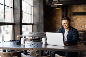Man sitting at a desk in a cafe with a laptop, smiling and looking at the camera.