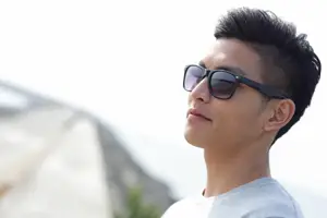 A man with a mohawk hairstyle wearing sunglasses and a white shirt looks up towards the sky in a sunny outdoor setting