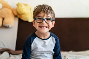 A young boy with glasses is smiling and sitting on a bed with stuffed toys behind him
