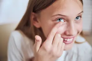 A young girl with brown hair wearing a white shirt and attempting to put a contact lens in her eye while smiling