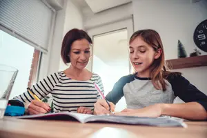 An adult woman and a girl are sitting in front of a table with a book and pens in a room