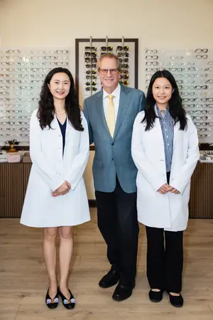 Three people are standing in front of a wall with many eyeglasses displayed on it. They are all smiling and seem to be posing for a photo.