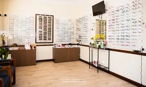 An interior of an optical shop with many eyeglasses displayed on the wall, a television mounted on the wall, and a small table with plants and flowers.