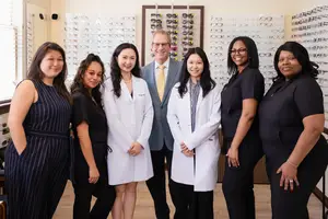 A group of women and a man in white coats and black dresses posing for a photo in a room with glasses on display.
