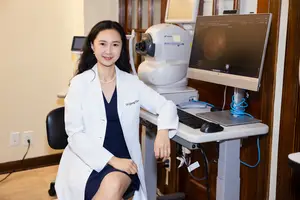 A female doctor wearing a white coat and a necklace is sitting in a chair in front of a desk with a monitor, keyboard, mouse, and medical equipment.