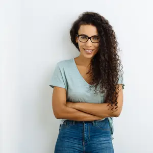 Portrait of a smiling woman with curly hair wearing glasses and a blue shirt, standing against a white background.