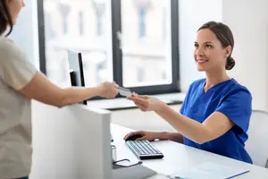 A smiling woman in a blue scrub is handing over a credit card to another woman in a tan shirt in an office setting