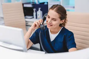 A woman in a blue shirt is sitting in an office and talking on the phone