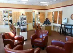 A modern waiting room with leather chairs, wooden tables, mirrors, and shelves with eyewear. The room features ceiling lights and a desk with a computer monitor.