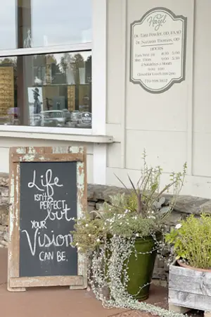 A vintage-style signboard with inspirational text and potted plants stands outside a building, with a reflection of a car and buildings in the glass window.