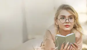 A woman wearing glasses and a pink jacket is reading a book on a pillow outside in the daytime.