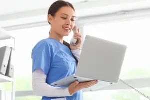 A woman in blue scrubs is smiling and looking at a laptop while holding a phone to her ear