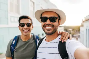 Two men in sunglasses and hats smile while posing for a selfie in front of a building.