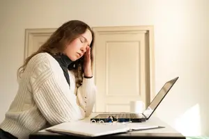 A woman with her head resting on her hand sitting at a desk with a laptop and cup.