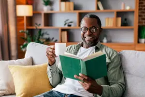 A man sitting on a couch reading a book while holding a cup of coffee