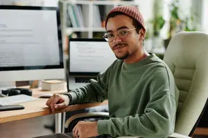 A man in a green shirt and glasses sits at a desk with a computer and a laptop on it.