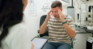 A man is sitting in a chair in an eye clinic and seems to be talking to a woman in front of him. The woman is holding a tablet, and a machine is placed on the right side of the man. Behind them is a wall with a chart with the text 'F P T O Z'. The man is wearing a bracelet and a watch.
